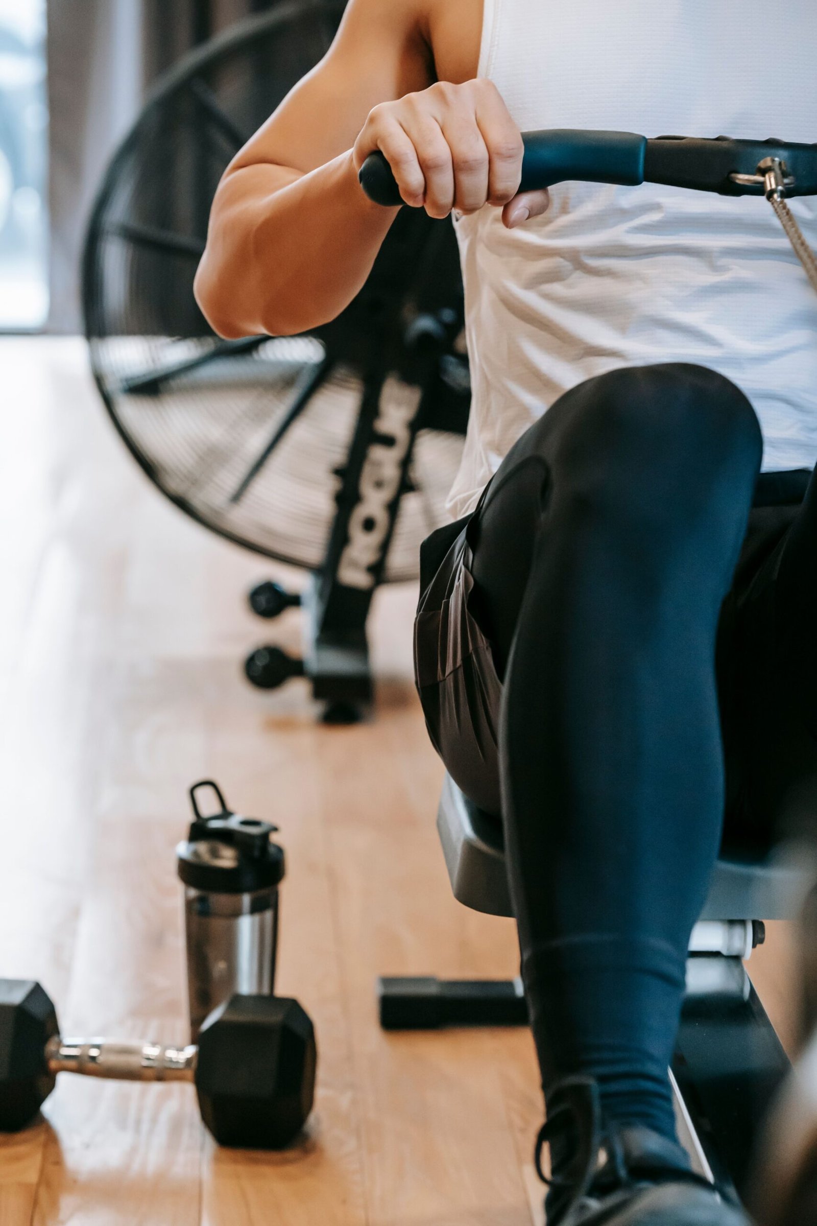 Crop anonymous male in sportswear exercising with rowing machine near dumbbell on floor